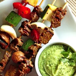 Steak and veggie skewers with a side of sauce on a white plate.