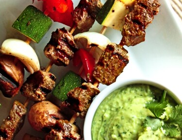 Steak and veggie skewers with a side of sauce on a white plate.