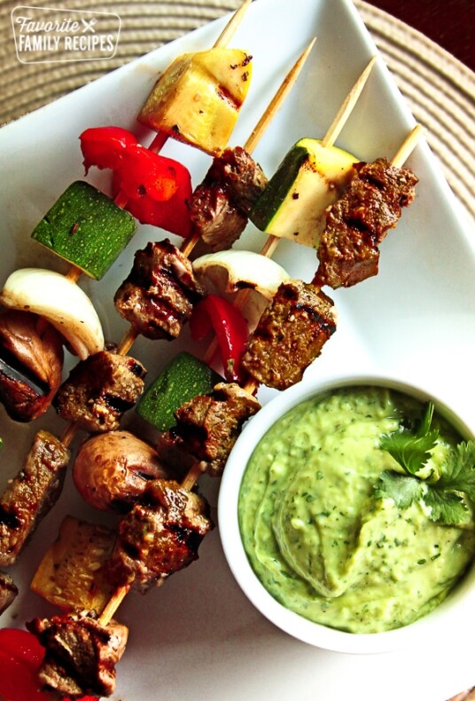 Steak and veggie skewers with a side of sauce on a white plate.