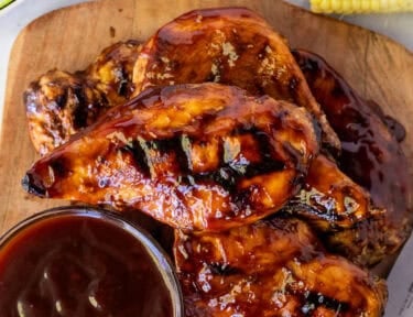 Grilled honey BBQ chicken breasts with char marks and glossy barbecue sauce on a wooden board beside extra sauce, corn, and watermelon.