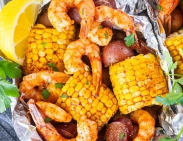 Cajun shrimp foil packets with corn on the cob, red potatoes, and sausage, garnished with parsley and lemon wedges.
