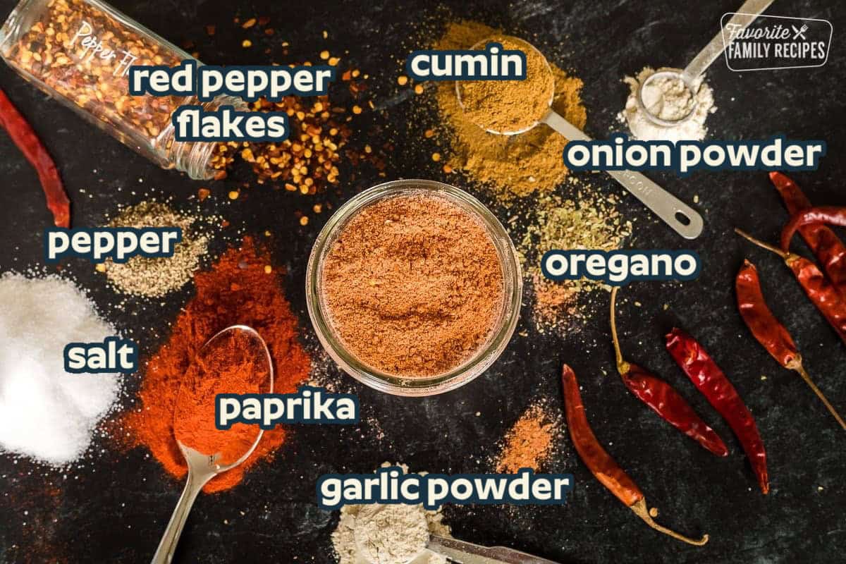Ingredients to make homemade ground beef taco seasoning in spoons and small piles on a black tabletop including pepper flakes, chili powder, cumin, onion powder, garlic powder, paprika, salt, and pepper.