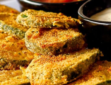 Baked fried zucchini on a white plate with stacked zucchini and marinara sauce.