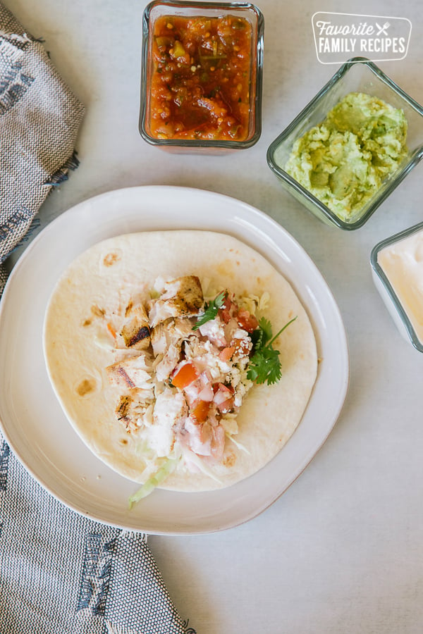 An open Grilled Mahi Mahi Fish Taco on a white plate with guacamole, salsa, and sour cream on the side