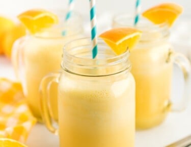 Three clear mugs full of orange julius with a blue straw and orange slice garnishes