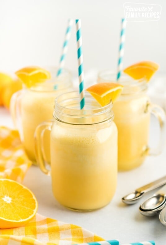 Three clear mugs full of orange julius with a blue straw and orange slice garnishes