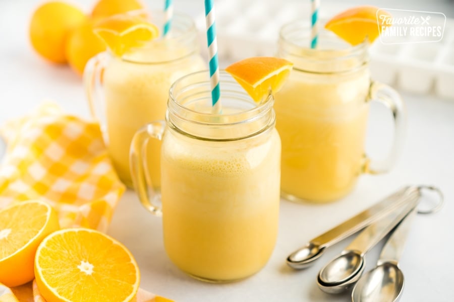 Three mugs of orange Julius with blue straws and orange slice garnishes.