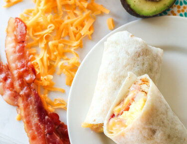 Breakfast burritos on a plate next to a block of cheese and an avocado