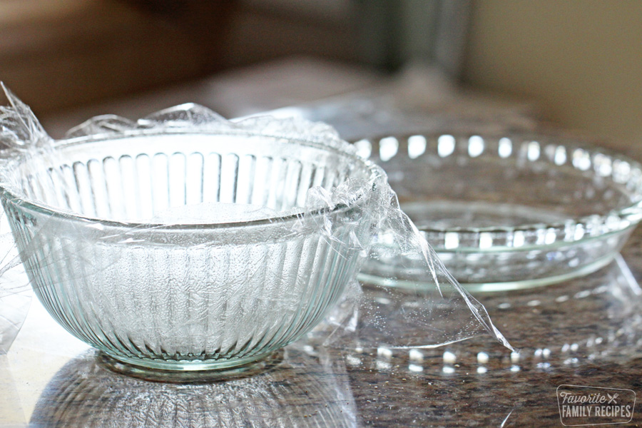 A glass mixing bowl lined with plastic wrap next to a glass pie plate