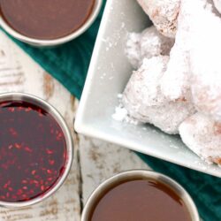 3 dipping sauces in little silver bowls next to a bowl of beignets covered in powdered sugar.