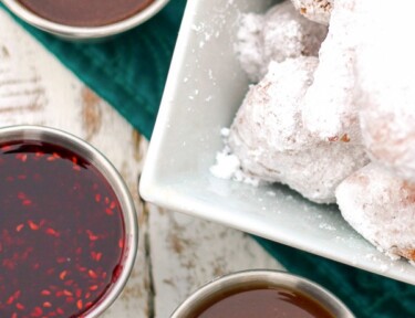 3 dipping sauces in little silver bowls next to a bowl of beignets covered in powdered sugar.