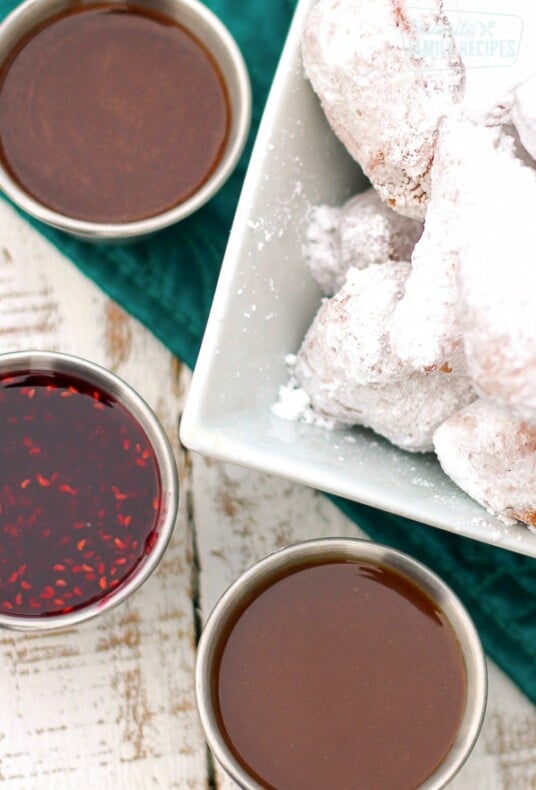 3 dipping sauces in little silver bowls next to a bowl of beignets covered in powdered sugar.