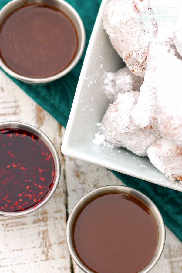 3 dipping sauces in little silver bowls next to a bowl of beignets covered in powdered sugar.