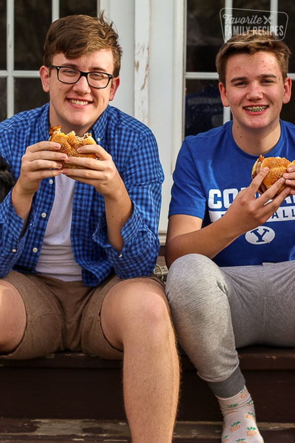 Two teenagers sitting on the back porch eating chicken sandwiches.