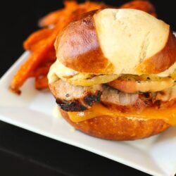 Grilled Pork Loin and Apple Burgers on a Pretzel Bun with sweet potato fries on the side on a white plate.
