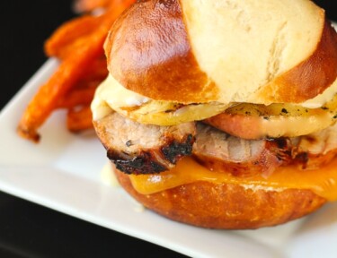 Grilled Pork Loin and Apple Burgers on a Pretzel Bun with sweet potato fries on the side on a white plate.