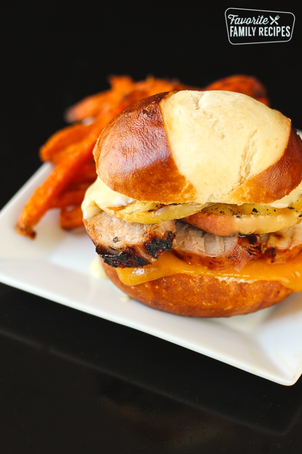 Grilled Pork Loin and Apple Burgers on a Pretzel Bun with sweet potato fries on the side on a white plate.