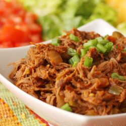 Crock Pot Mexican Shredded Beef served in a white bowl on a colorful placemat with chopped tomatoes, lettuce, and shredded cheese in the background
