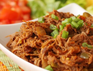 Crock Pot Mexican Shredded Beef served in a white bowl on a colorful placemat with chopped tomatoes, lettuce, and shredded cheese in the background