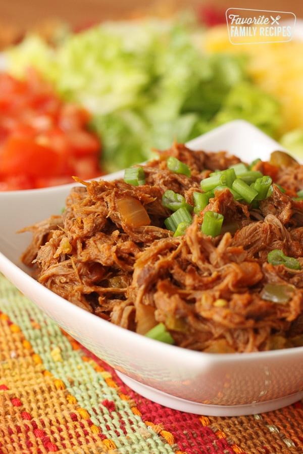 Crock Pot Mexican Shredded Beef served in a white bowl on a colorful placemat with chopped tomatoes, lettuce, and shredded cheese in the background.