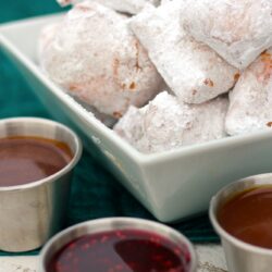 New Orleans Mini Beignets covered in powdered sugar with three dipping sauces on the side.