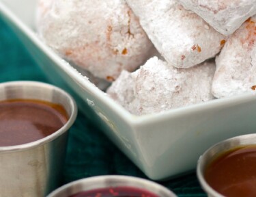 New Orleans Mini Beignets covered in powdered sugar with three dipping sauces on the side.