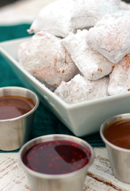 New Orleans Mini Beignets covered in powdered sugar with three dipping sauces on the side.