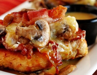 Outback's Alice Springs Chicken topped with bacon and mushrooms on a white tray with a side of sauce.