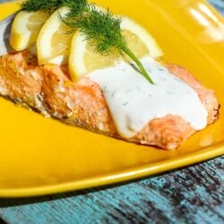 Baked Lemon Salmon covered Tzatziki Sauce,3 lemon slices, and parsley on a yellow plate.