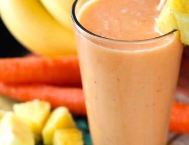 Golden Detox Smoothie in a tall glass with pineapples, bananas, and carrots on the side.