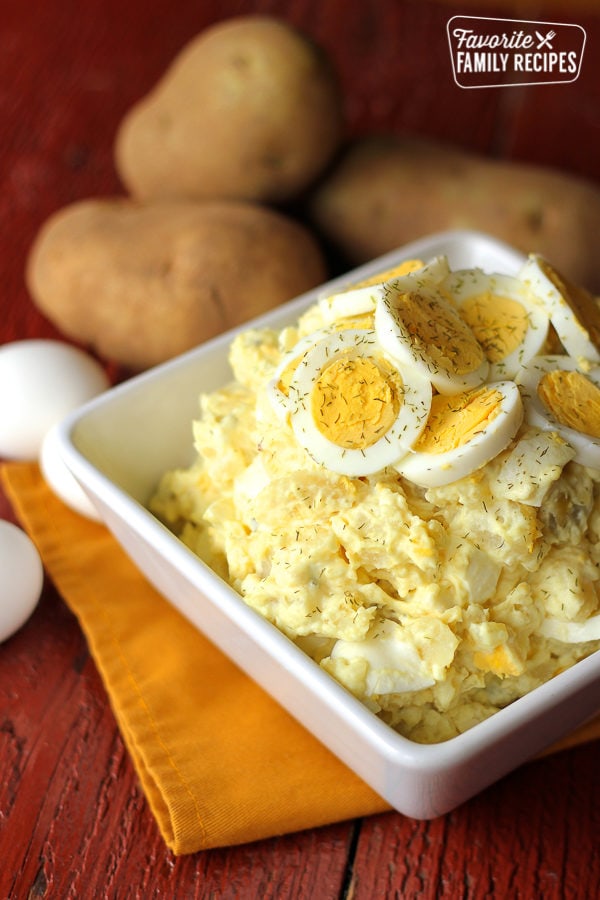 potato salad with egg slices on top in a white bowl and potatoes in background