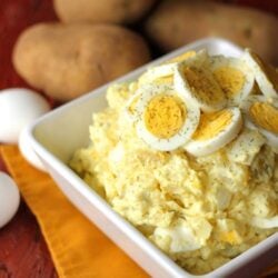 potato salad with egg slices on top in a white bowl and potatoes in background