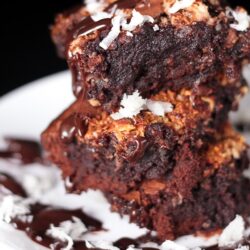 3 almond joy brownies stacked on top of each other covered in chocolate sauce and coconut flakes on a plate.