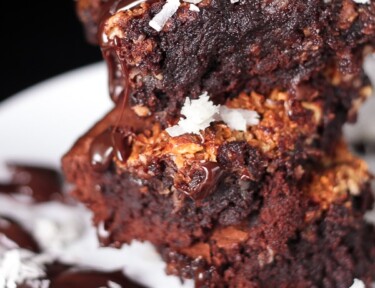 3 almond joy brownies stacked on top of each other covered in chocolate sauce and coconut flakes on a plate.