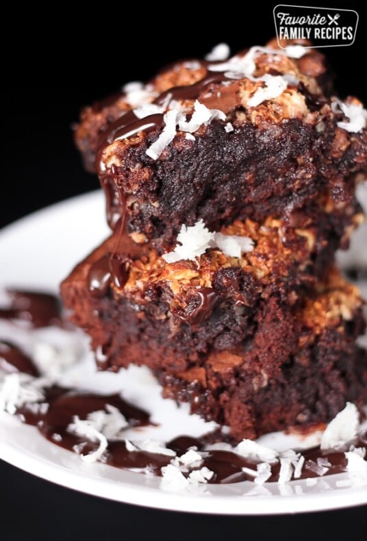 3 almond joy brownies stacked on top of each other covered in chocolate sauce and coconut flakes on a plate.