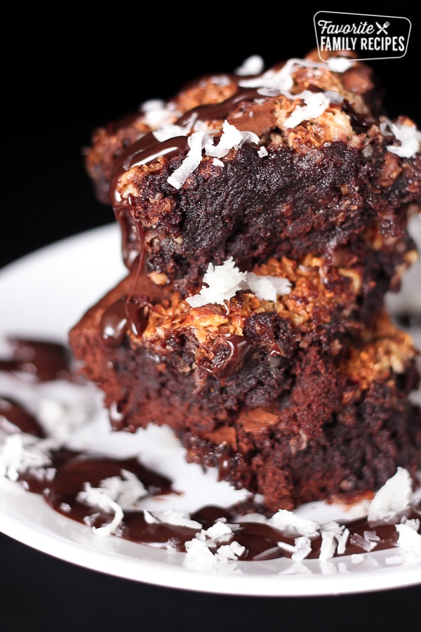 3 almond joy brownies stacked on top of each other covered in chocolate sauce and coconut flakes on a plate.