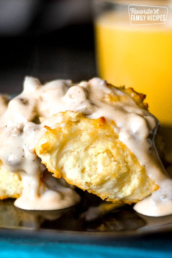 2 Biscuits covered Sausage Gravy on a plate with a glass of orange juice in the background.