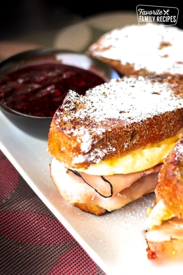 Breakfast Monte Cristos on a plate covered in powdered sugar with a bowl of jam in the background