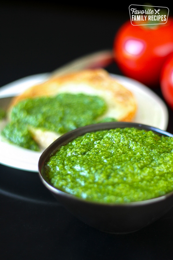 Fresh Homemade Basil Pesto in a black bowl with more pesto and tomatoes in the background.