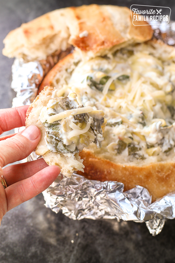 spinach artichoke dip in a bread bowl with piece of bread in hand