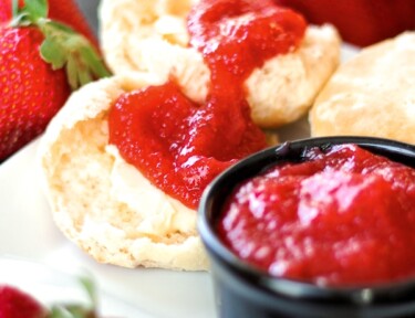 Fresh and Easy Strawberry Jam in a small black bowl with rolls and jam on the side on a white plate surrounded by strawberries.