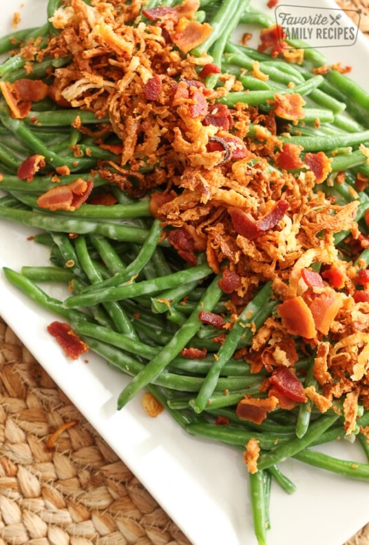 Fresh green beans on a white platter topped with bacon bits and onion straws