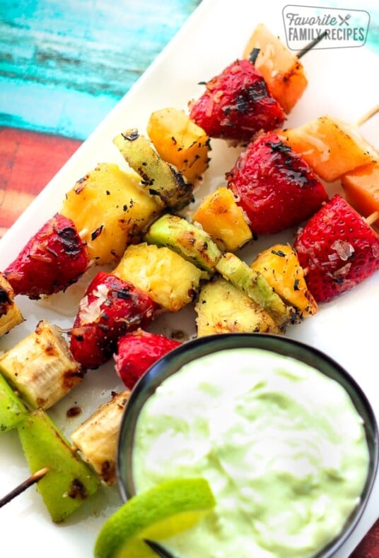 3 Grilled Fruit Kabobs with Key Lime Yogurt Dip on othe side all on a white plate.