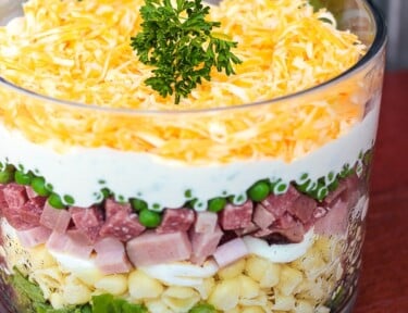 Layered Pasta Salad with ham, peas, noodles in a glass dish topped with cheese and a garnsih.