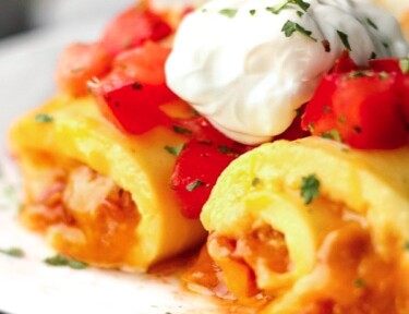 Omelette Roll Ups topped with sour cream and tomatoes on a white plate.