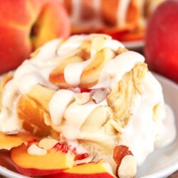 Peaches and Cream Cinnamon Roll with a couple peach slices on the side on a white plate.