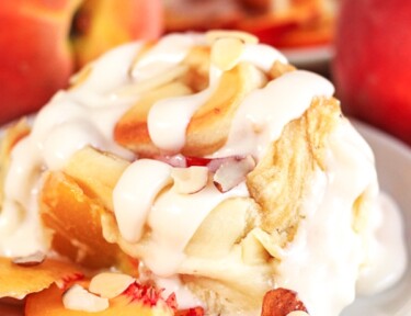 Peaches and Cream Cinnamon Roll with a couple peach slices on the side on a white plate.