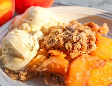 Crock Pot Peach Cobbler served with two scoops of vanilla ice cream with fresh peaches in the background