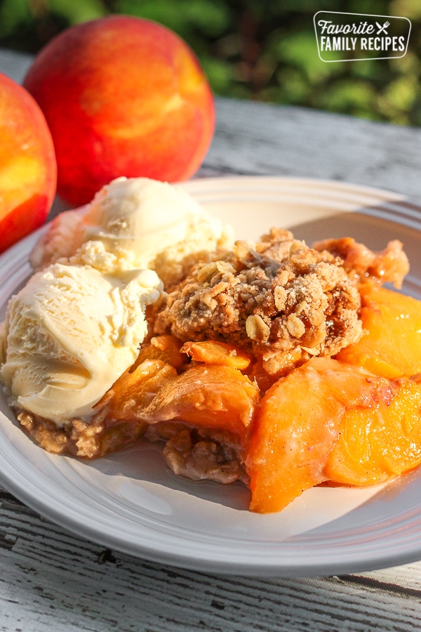 Crock Pot Peach Cobbler served with two scoops of vanilla ice cream with fresh peaches in the background.