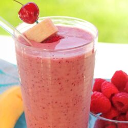 Banana berry smoothie in a glass with a cherry and banana slice on a toothpick and a bowl of raspberries in the background